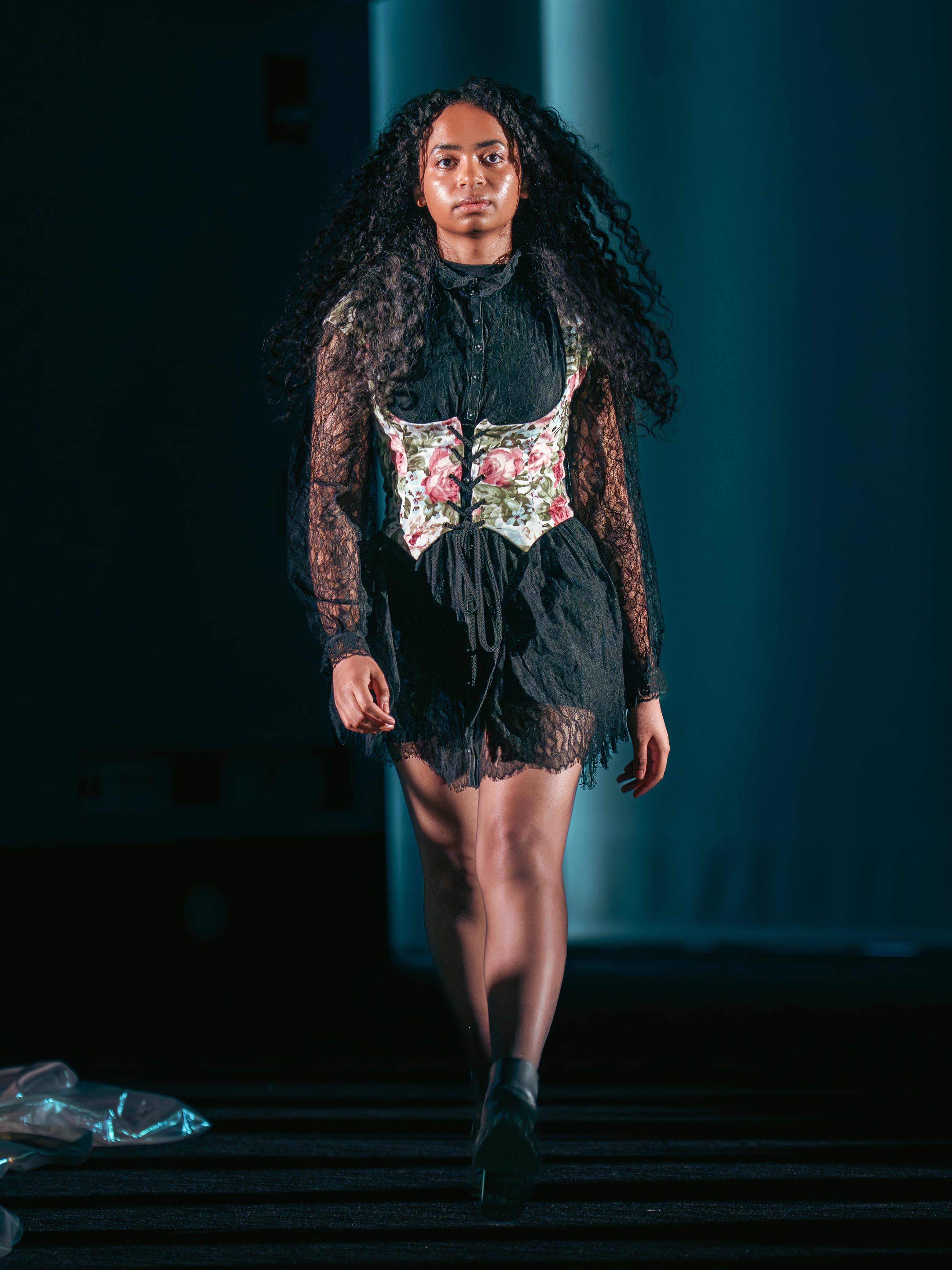 Model walks in a black lace dress with long sleeves and a floral corset overlay. The short hemline reveals sheer tights and ankle boots, with voluminous curly hair framing her face.